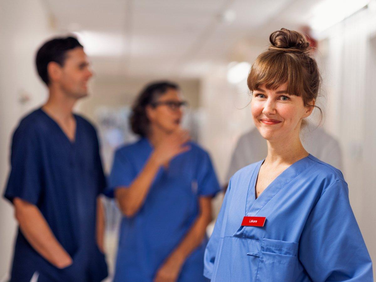 En ung kvinnlig läkare som står i en korridor i sjukhusmiljö. Hon tittar in i kameran och ler. I bakgrunden står tre kollegor till läkaren och pratar med varandra.